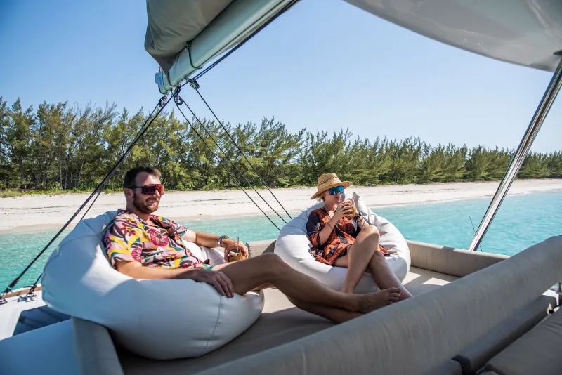 Two people on white bean bags on deck with coconut drinks, a white sand beach behind them