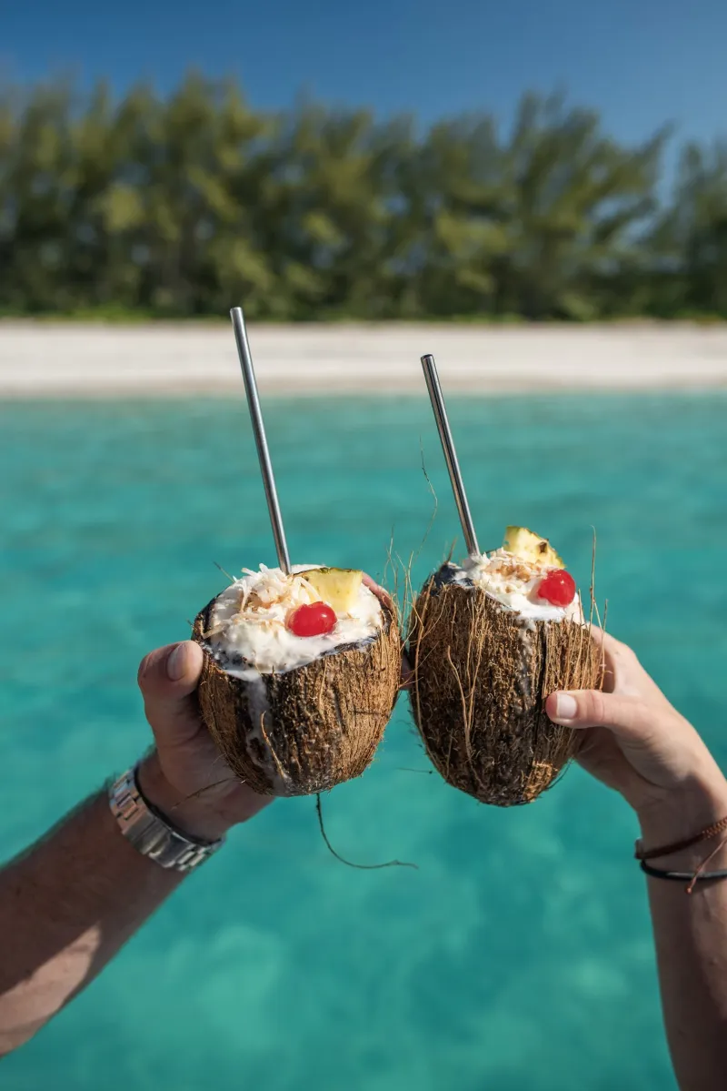 Two coconut cocktails topped with pineapple and a cherry, held up over turquoise water