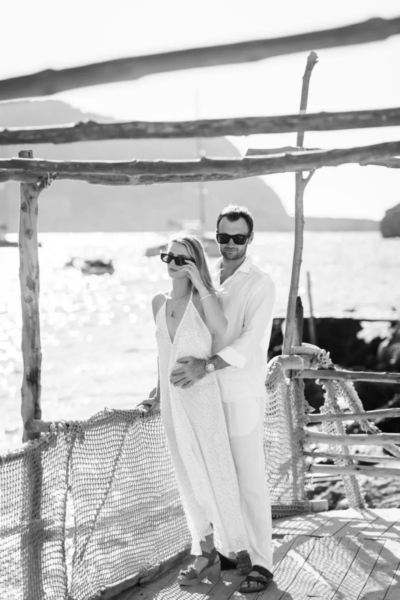 Black and white photo of Lily and Nik in sunglasses under a driftwood pergola by the sea